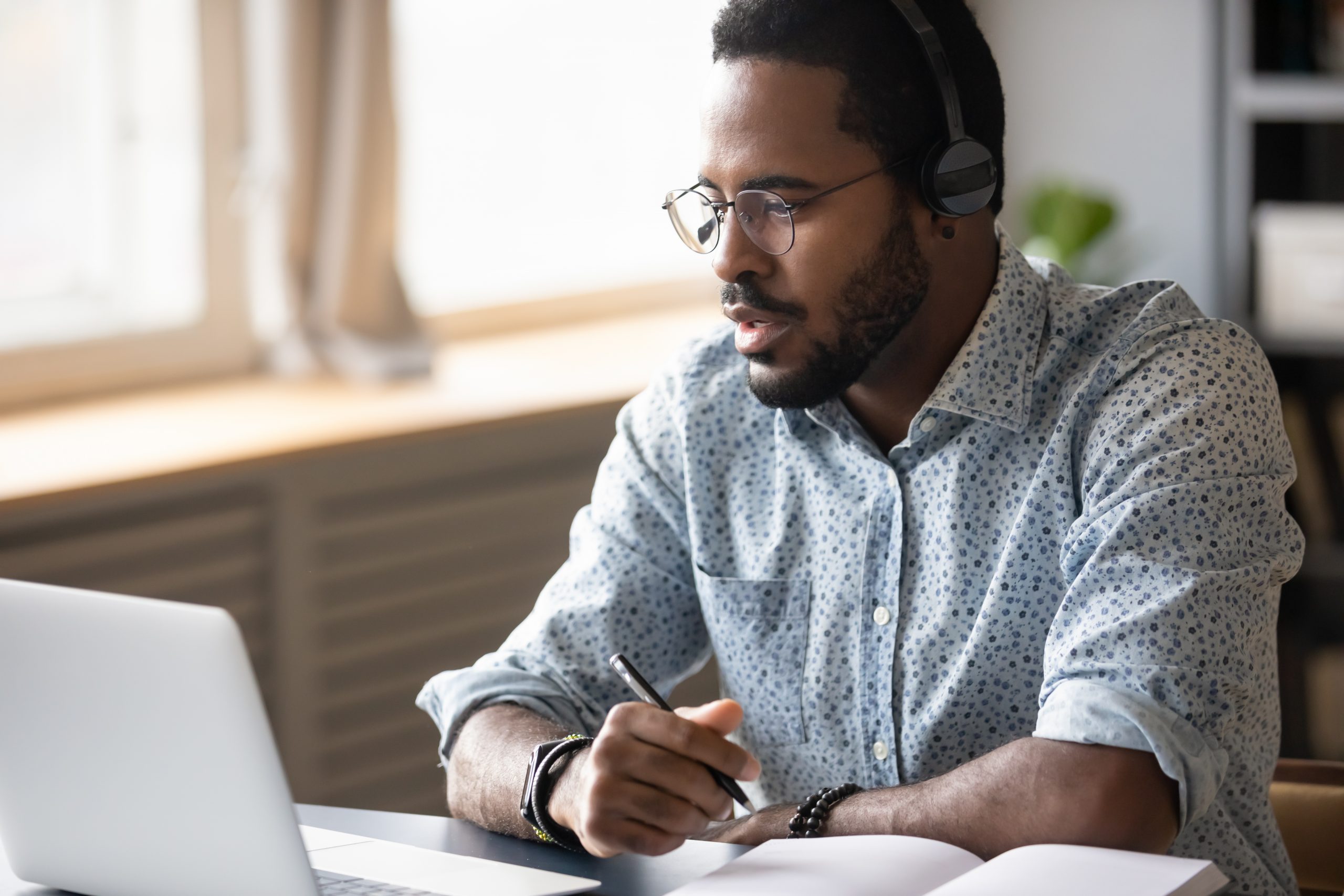 Head shot focused young african american man in glasses wearing wireless headphones, looking at laptop screen, watching educational lecture seminar webinar online, studying remotely, writing notes.