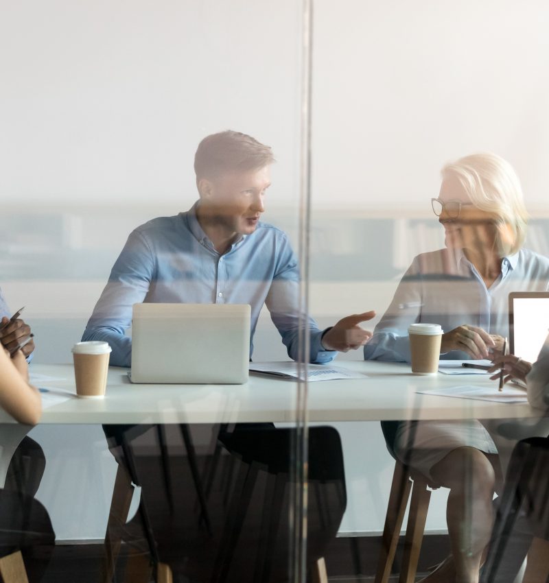 Diverse business people negotiate sit at conference table behind closed glass door at group private meeting, multi ethnic team staff employees talk at briefing discuss work in modern office boardroom
